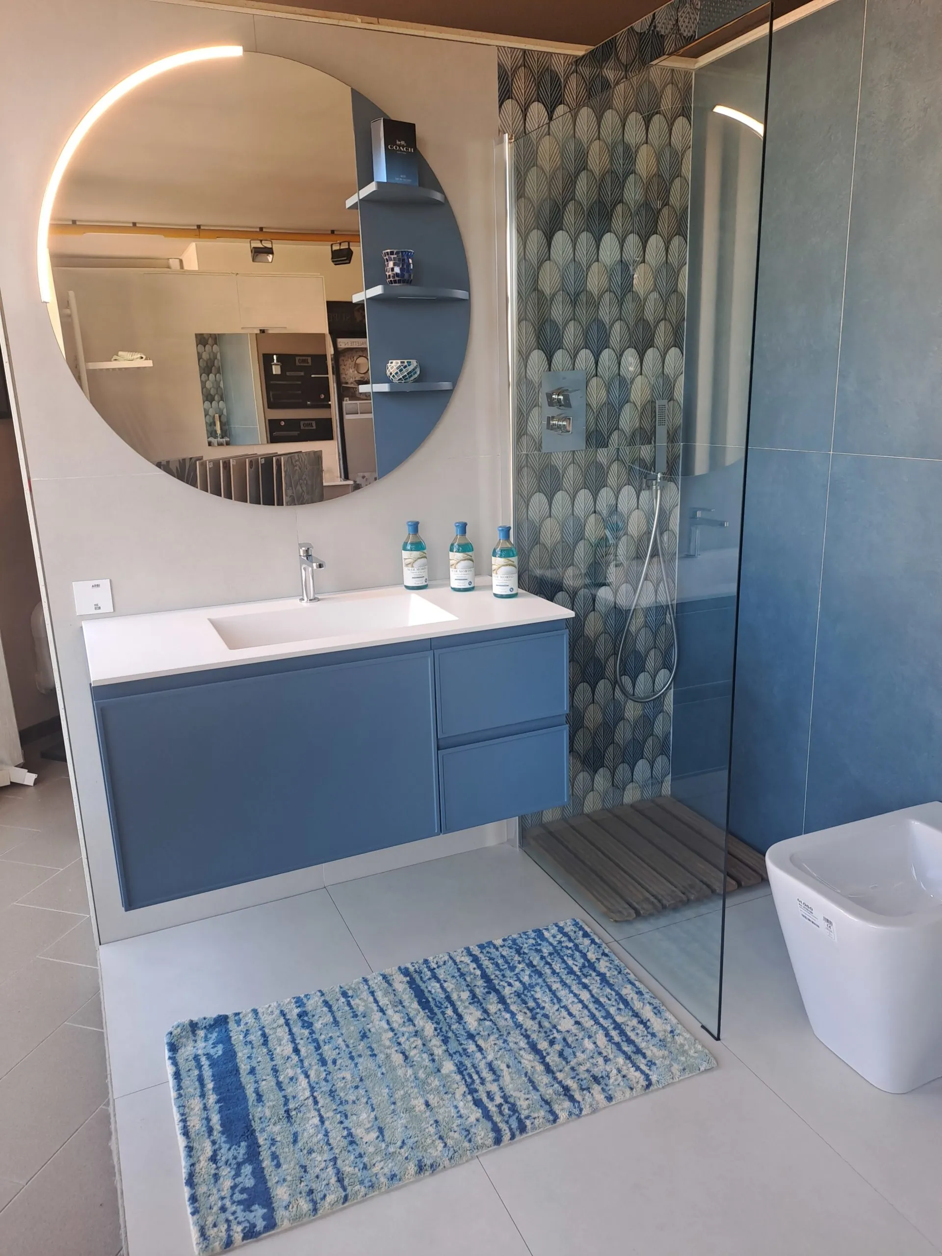 Bagno con mobile sospeso blu, grande specchio tondo e doccia con rivestimento a scaglie